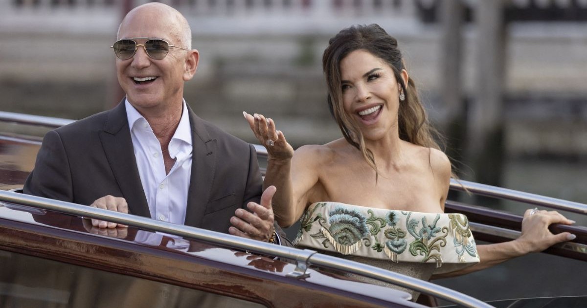 Jeff Bezos, in sacou gri, camasă albă si ochelari de soare, și Lauren Sanchez, intr-o rochie fara umeri cu model brodat și cu mana dreaptă ridicată, părăsesc hotelul - Veneția, Italia - 26 iunie 2025