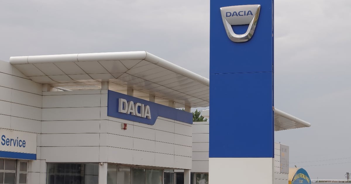 Imaginea surprinde un showroom Dacia, cu logo-ul brandului vizibil pe clădire și pe un panou înalt amplasat în față. Spațiul pare modern și bine organizat, sugerând un punct de vânzare și service auto.