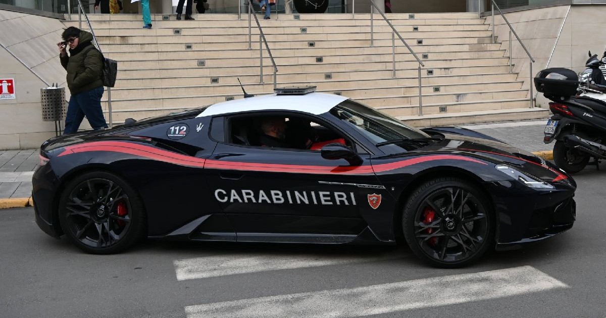 Fotografie cu un Maserati al carabinierilor parcat pe o stradă, în fața scărilor unei clădiri, în timp ce un bărbat îmbrăcat într-o jachetă verde trece pe lângă mașină.