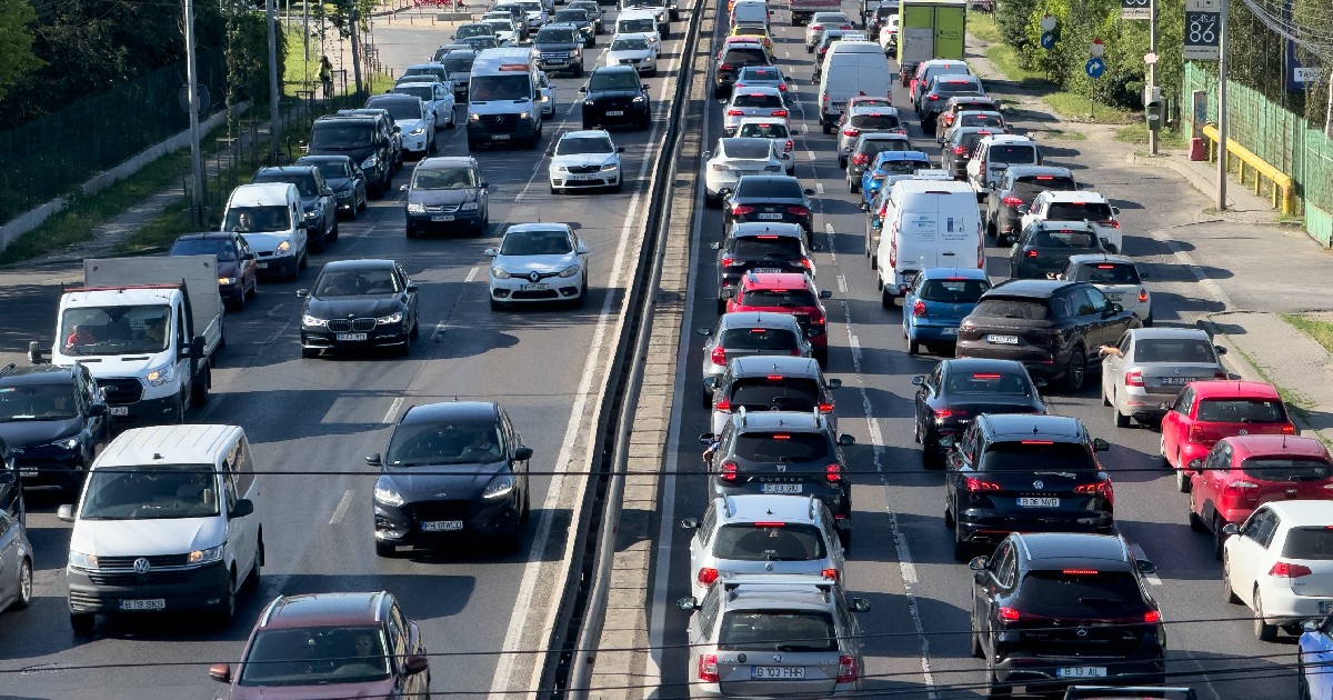 Imaginea prezintă un trafic rutier intens pe o arteră cu mai multe benzi, unde numeroase autoturisme și utilitare sunt surprinse circulând bară la bară.