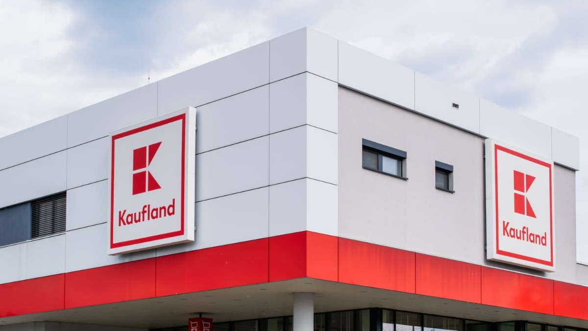 În imagine este surprinsă fațada unui magazin Kaufland. Clădirea are un design modern, cu panouri albe și gri, iar în partea superioară se află logo-ul Kaufland pe fundal roșu, afișat de două ori.