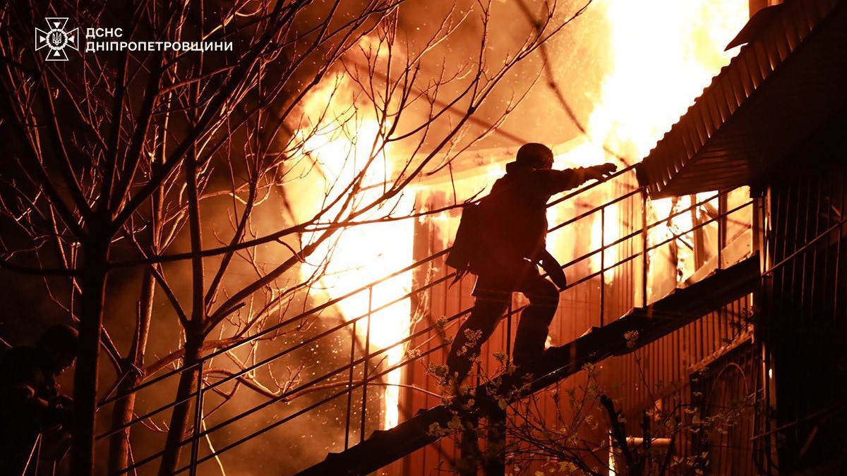 Un salvator ucrainean urcă pe o scară pentru a ajunge la o clădire din Dnipro. Lângă el, o clădire este cuprinsă de flăcări
