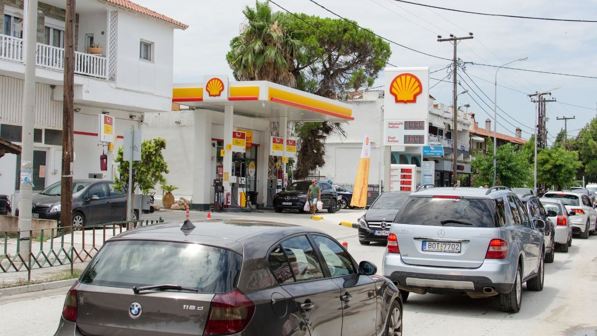 Imaginea surprinsă în Halkidiki, Grecia, prezintă o stradă intens circulată, mărginită de clădiri cu etaj și vegetație bogată, printre care se distinge trunchiul și coroana unui palmier. În prim-plan, carosabilul este ocupat de o coloană compactă de autoturisme oprite în trafic, ieșind în evidență o mașină cu formă rotunjită în partea stângă și un vehicul de teren în dreapta. Pe partea stângă a străzii, integrată în peisajul urban, se află o stație de alimentare cu carburanți. Aceasta este alcătuită dintr-un acoperiș plat susținut de stâlpi groși, sub care sunt amplasate pompele de alimentare, și un panou informativ foarte înalt pe care tronează o siglă în formă de scoică și lista de prețuri. În spațiul benzinăriei sunt parcate alte vehicule, iar un pieton este surprins traversând zona pompelor, în timp ce de-a lungul întregii străzi se înșiruie stâlpi de utilități interconectați prin cabluri aeriene.