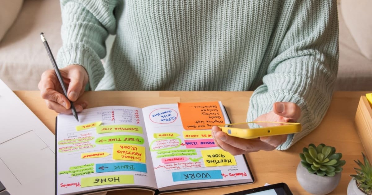 Femeie imbracata intr-un pulover verde menta sta la birou si isi organizeaza timpul intr-o agenda. Intr-o mana tine un pix cu care scrie, in cealalta telfonul, iar in fata are o tableta