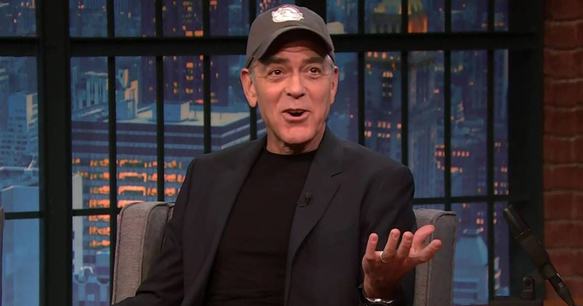 George Clooney în timpul unei emisiuni TV, stă pe canapea într-un costum negru, gesticulează cu mâinile și poartă o șapcă pe cap.
