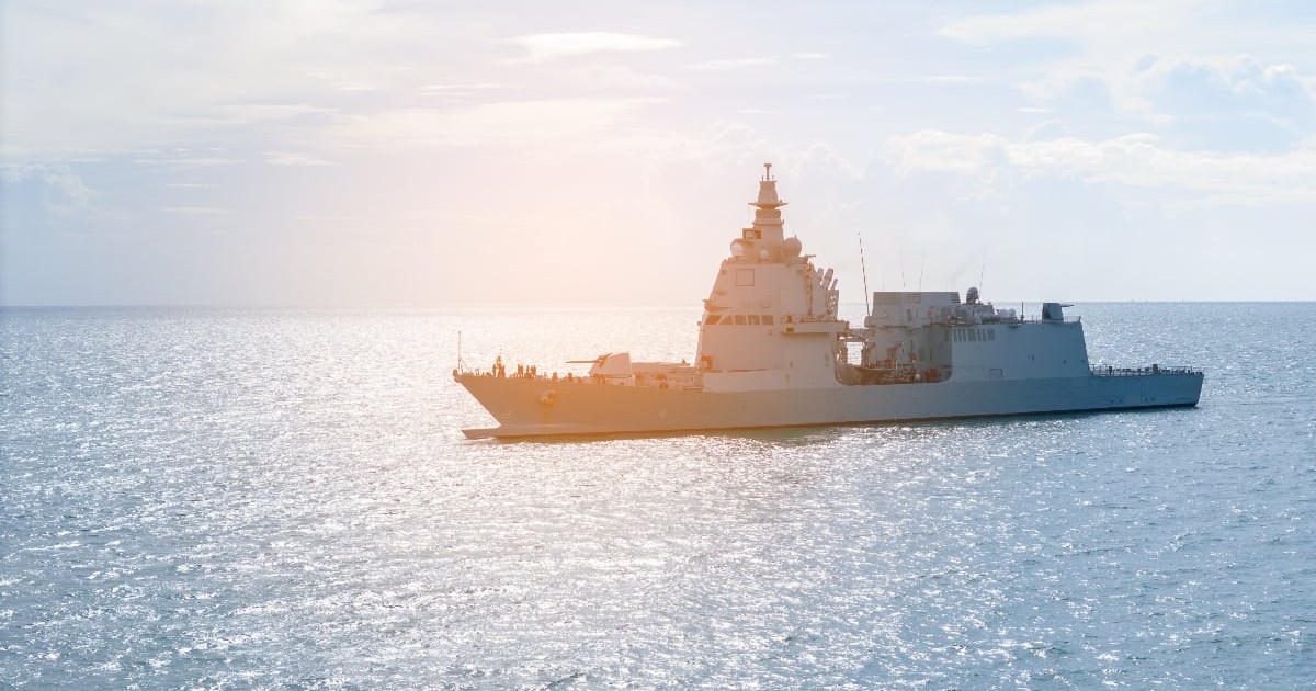 Imaginea surprinde o navă militară de mari dimensiuni navigând pe mare, în condiții de vreme senină, cu soarele reflectându-se pe suprafața apei. Nava este văzută din lateral, fără semne vizibile de avarii, într-un cadru care sugerează o misiune de patrulare sau exercițiu naval.