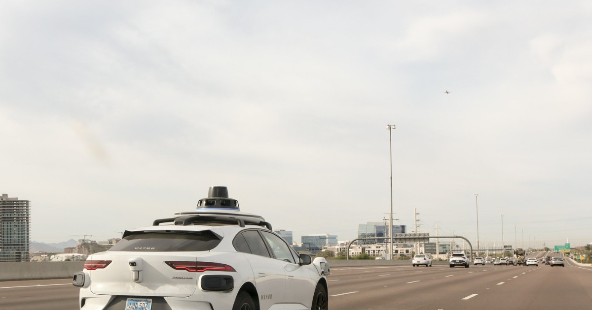 Imaginea prezintă o mașină autonomă Waymo care circulă pe o autostradă largă, alături de alte vehicule, într-un peisaj urban modern. Pe plafonul automobilului se observă senzorii și echipamentele specifice conducerii autonome, iar în fundal se disting clădiri și infrastructură specifică unei zone metropolitane.