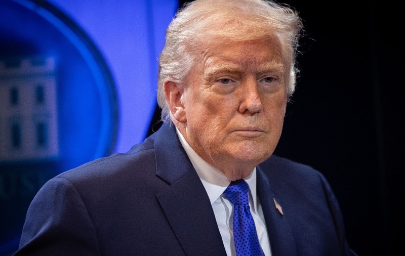 Președintele american Donald Trump îmbrăcat în costum închis la culoare și cravată albastră, privind serios în cadrul unei conferințe oficiale.