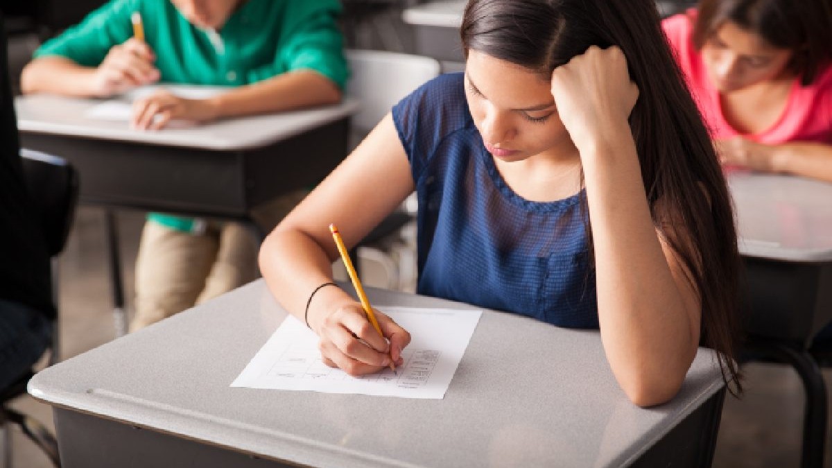 O fata imbracata intr-o bluza bleumarin cu maneca scurta sta intr-o banca si scrie. Canicula la examen.