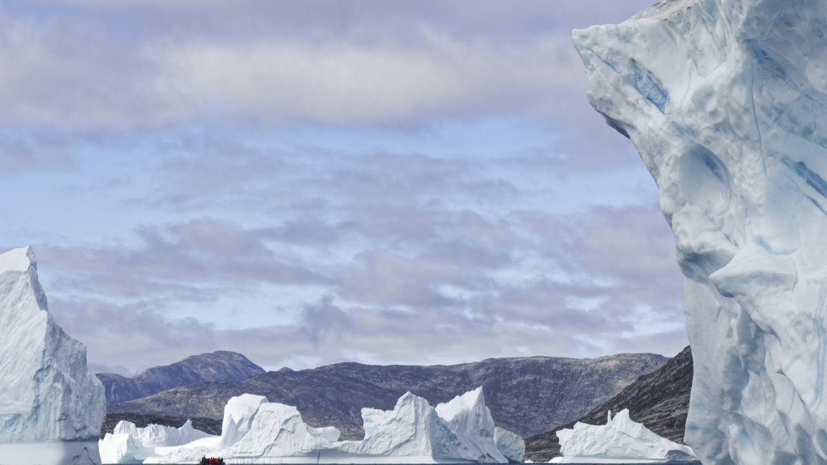 Imaginea surprinde un peisaj arctic din Groenlanda, cu ghețari masivi care plutesc pe o mare întunecată, având în fundal munți stâncoși. Atmosfera rece și sălbatică evidențiază fragilitatea naturii și izolarea extremă a regiunii.