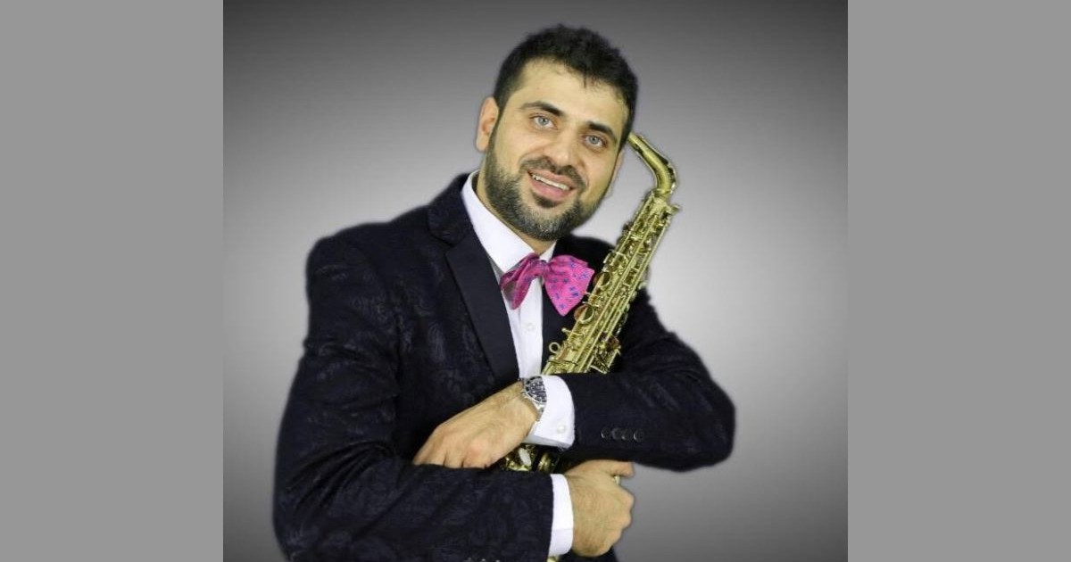 Memiș Selciuc, îmbrăcat într-un costum negru, cu un papion roz la gât și un saxofon auriu în brațe