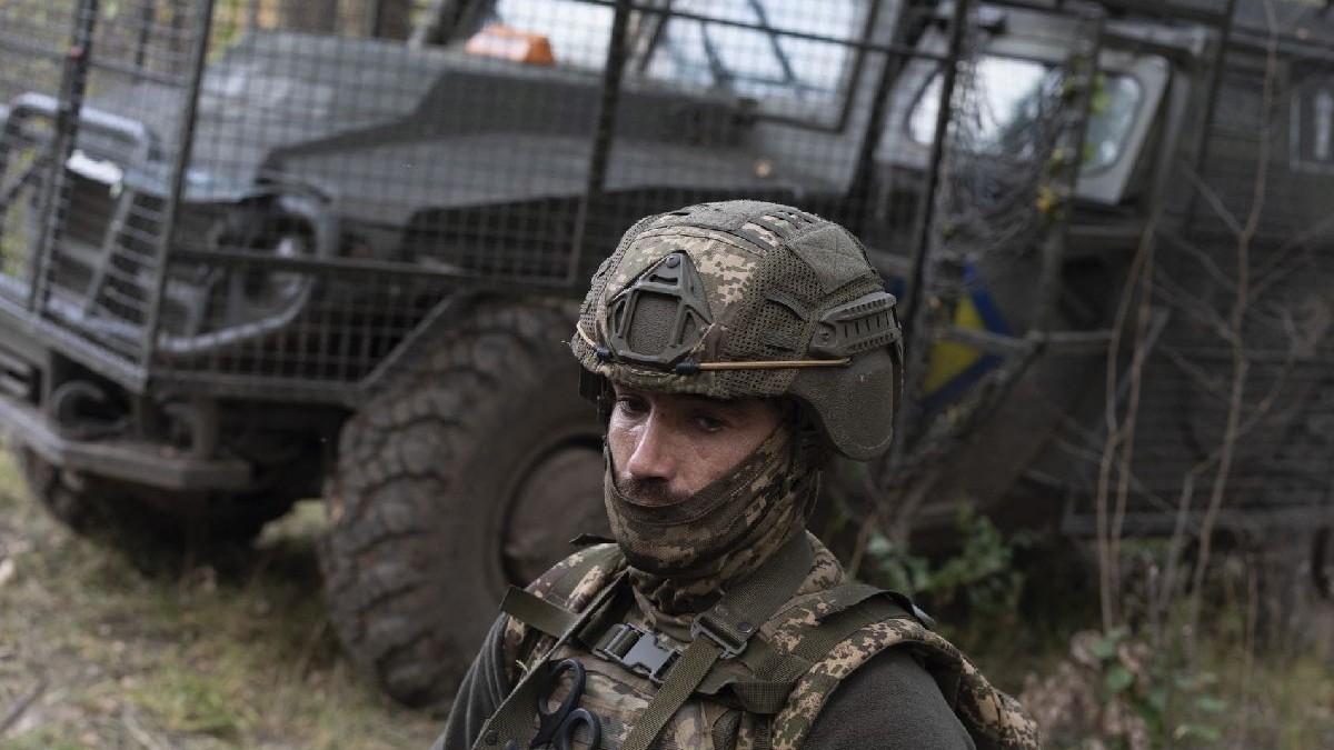 Un soldat în echipament de camuflaj și cască stă într-o zonă împădurită, cu un gard și un vehicul militar în fundal.
