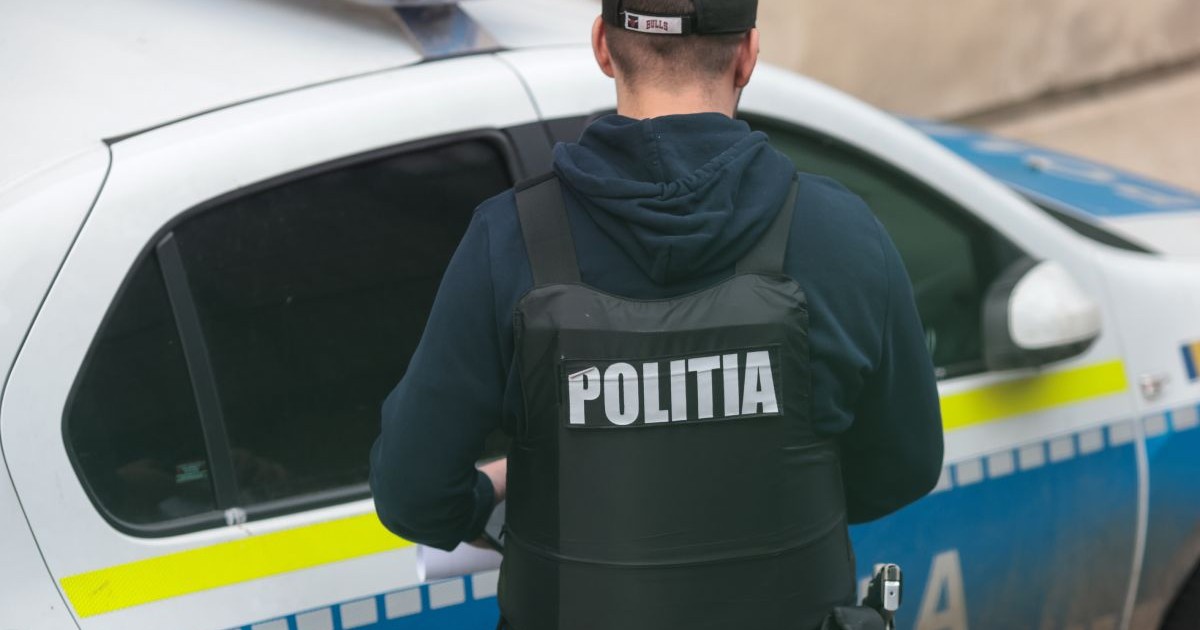 Polițist îmbrăcat cu o vestă pe care scrie Poliția, fotografiat din spate, lângă o mașină de Poliție