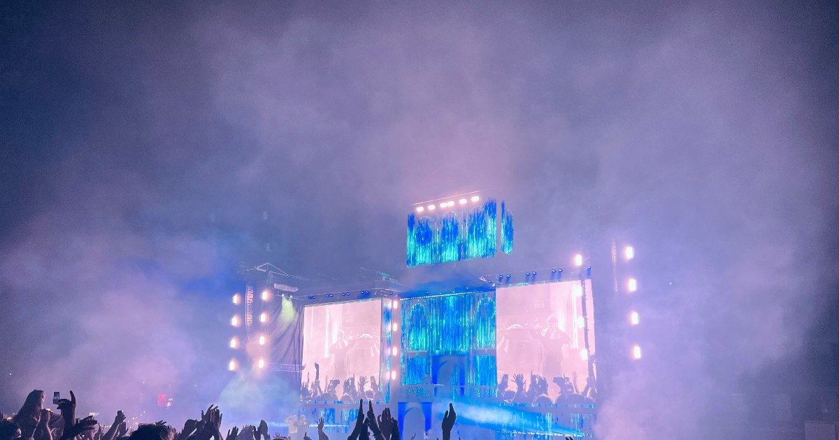 Imaginea surprinde un concert în aer liber, cu o scenă mare iluminată și ecrane uriașe, în fața unei mulțimi numeroase care ridică mâinile și filmează momentul. Atmosfera este una energică, accentuată de lumini, fum de scenă și entuziasmul publicului.