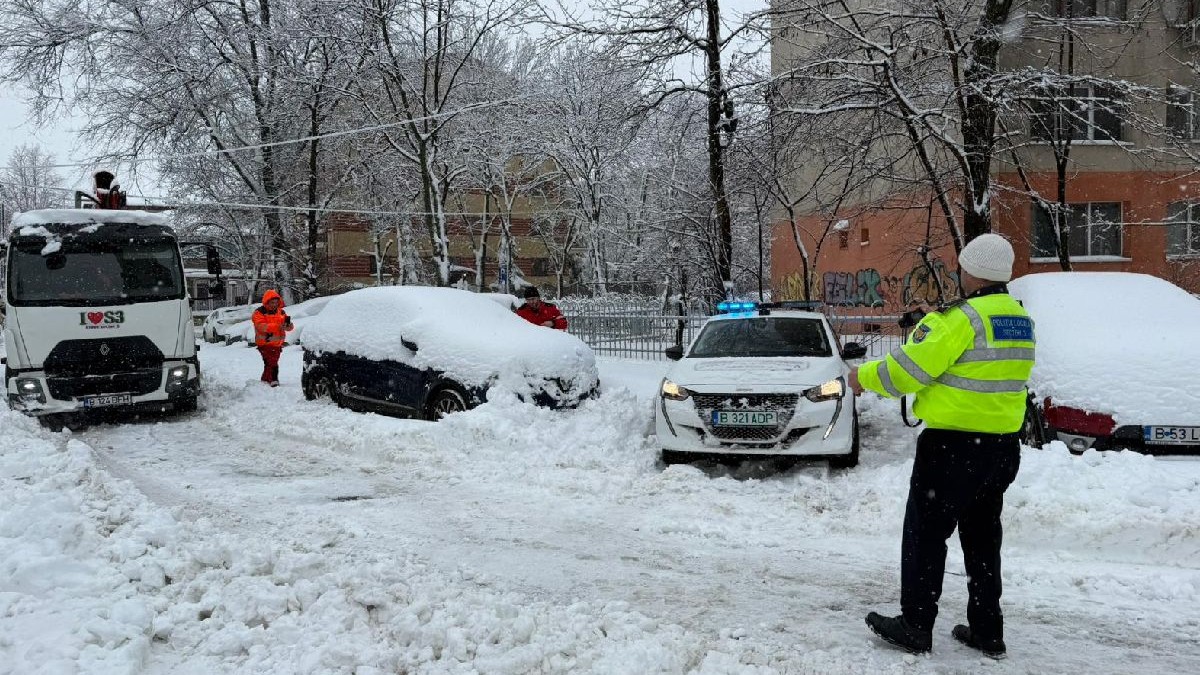 Pe o stradă acoperită de zăpadă, un polițist supraveghează o mașină care ridică autoturisme parcate ilegal, în București
