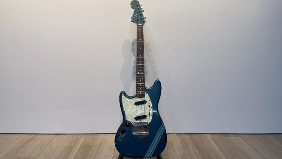 În imagine se vede o chitară electrică albastră, expusă pe un suport, cu un design clasic și pickguard deschis la culoare. Instrumentul este fotografiat într-un spațiu simplu, cu perete alb și podea din lemn, punând accent pe forma și detaliile chitarei.