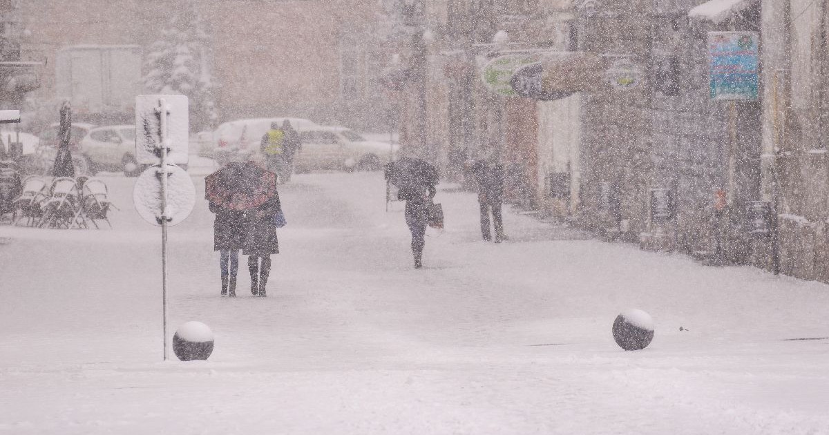 Fotografie cu o stradă înzăpezită pe care patru persoane îmbrăcate în haine de iarnă se îndepărtează. Clădiri acoperite de zăpadă de-a lungul ambelor părți ale străzii, iar câteva mașini parcate sunt vizibile. Ninsoare ușoară, o atmosferă liniștită, de iarnă.
