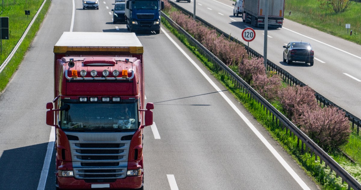 Vedere de sus a unei autostrăzi, înfățișând în prim-plan un camion de culoare roșie care circulă pe prima bandă, în timp ce pe sensul opus și în fundal se deplasează alte autovehicule