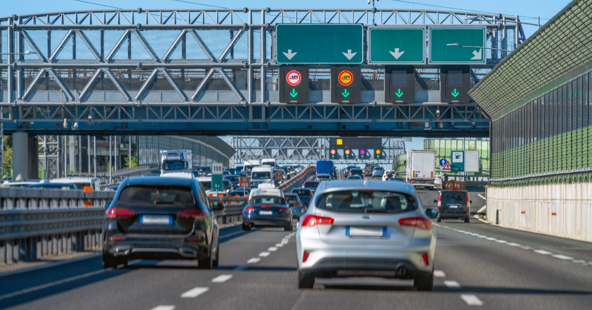 Imaginea prezintă un trafic auto intens pe o autostradă cu mai multe benzi, unde vehiculele se îndreaptă spre o pasarelă metalică masivă dotată cu panouri electronice de informare și semnalizare rutieră.