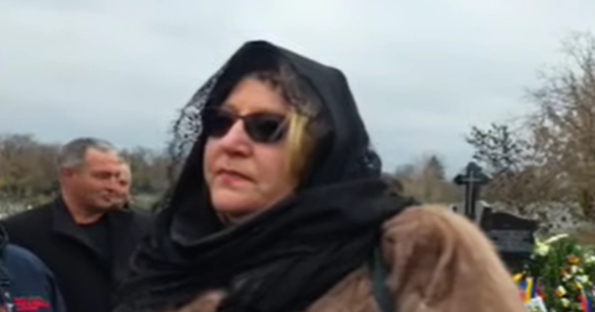 Femeia, identificată ca fiind Diana Șoșoacă, poartă o haină de blană maro și o eșarfă neagră pe cap, acoperindu-și parțial fruntea. Poza pare să fie făcută în aer liber, într-un cimitir, având în fundal câțiva oameni și o coroană funerară