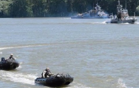 Un exercițiu militar pe apă, în care mai multe bărci pneumatice rapide ghidate de militari înaintează pe un fluviu, având nave militare de patrulare în fundal.