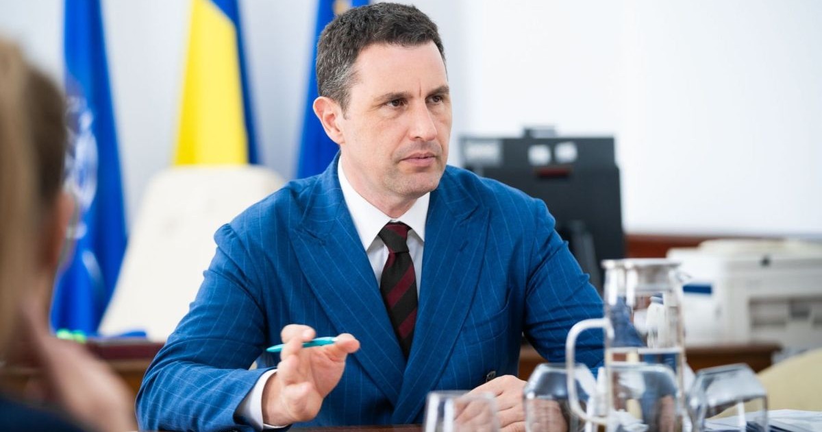 Ministrul interimar al Finanțelor, Tanczos Barna, îmbrăcat la costum, stă la o masă unde vorbește și explică dând din mâna stângă, în care are și un pix, iar în fața lui sunt pahare cu apă și în spate se văd steagul României, UE și al NATO
