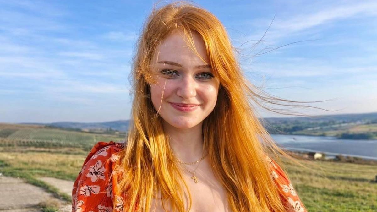 Andreea Anita, cu parul portocaliu, desfacut, zburat de vant, intr-o rochie rosie cu flori albe, zambeste intr-o zi insorita, se vede un cer albastru, in spate dealuri verzi și o apa, Andreea, din Falticeni, are nevoie de 2.3 milioane de dolari pt un tratament in SUA