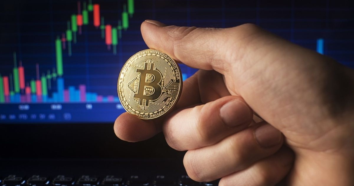 Un bărbat ține în mână o monedă bitcoin auriu pe fundalul graficului de tranzacționare pe piața bursieră