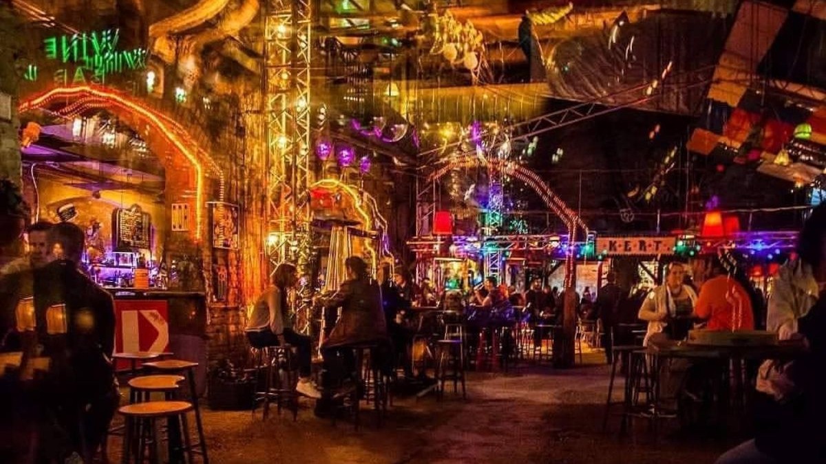 nteriorul unui local aglomerat, amenajat într-un spațiu vast cu un aspect brut, industrial, definit de pereți cu textură nefinisată, arcade ample și stâlpi înalți din schele metalice. Încăperea este decorată abundent și aparent haotic cu o multitudine de obiecte suspendate de tavanul înalt, incluzând globuri disco, lămpi cu abajururi de forme diferite, cabluri la vedere și diverse alte decorațiuni eclectice.