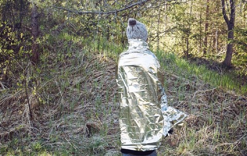 Imagine cu o femeie care s-a rătăcit și se află într-o pădure sălbatică, într-o zi rece, folosind o folie izolantă pentru a preveni hipotermia și pierderea căldurii corporale.