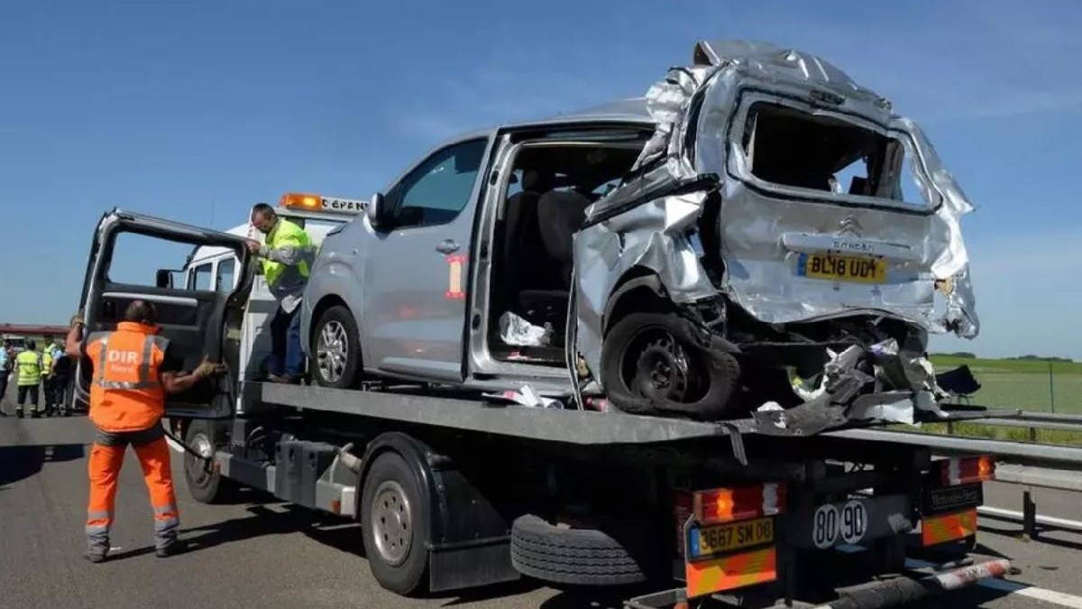 O fotografie cu o mașină argintie pe o platformă de remorcare după un accident, cu un bărbat îmbrăcat într-o jachetă portocalie care dirijează camionul.
