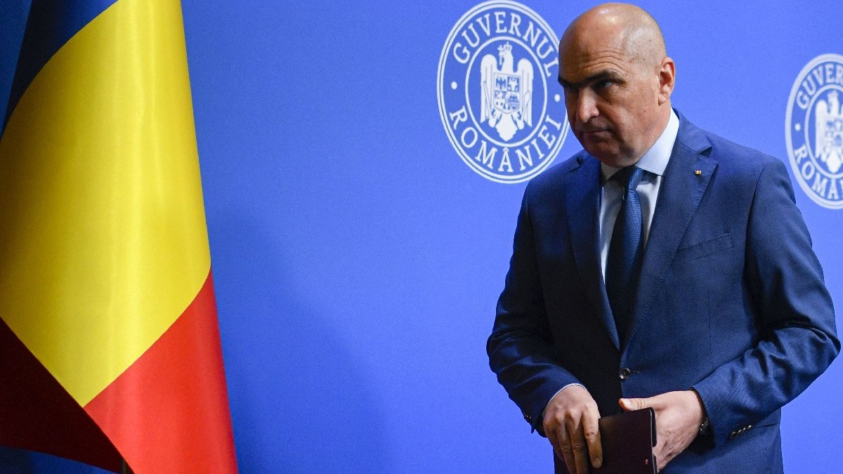 Premierul Ilie Bolojan, îmbrăcat în costum albadstru închis, pleaca la finalul unei conferinte de presa cu privire la reforma pensiilor in sitemul de Jutitie, marti, 29 iulie 2025, in Bucuresti. In fundal, în stânga se vede și steagul României.