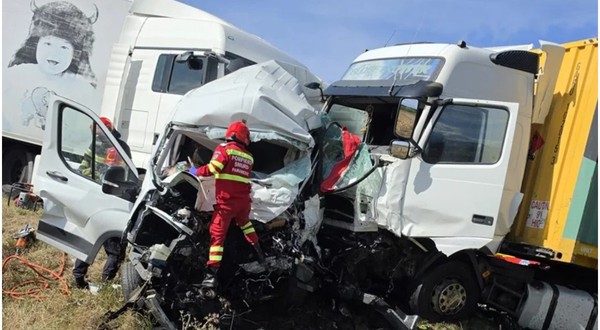 Imaginea prezintă urmările unui accident grav, în care o dubă albă este zdrobită de două TIR-uri, în timp ce un echipaj de pompieri și paramedici SMURD intervine la fața locului.
