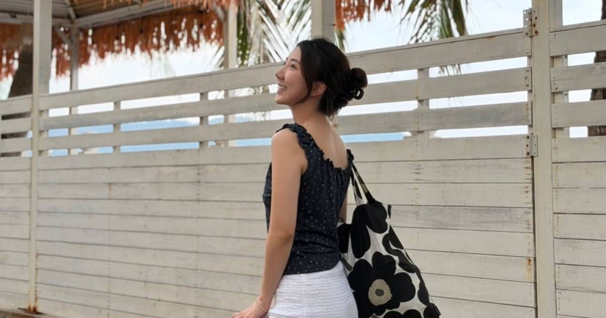 Luby, tânăra din Hong Kong care conduce camioane de mare tonaj și câștigă peste 3.200 de euro pe lună, îmbrăcată cu un top negru și o fustă albă, stă pe o terasă și privește zâmbind în lateral. Are părul prins într-un coc și poartă pe umăr o geantă mare, alb-negru, cu imprimeu floral. În fundal se văd un gard alb din lemn, palmieri și marea.