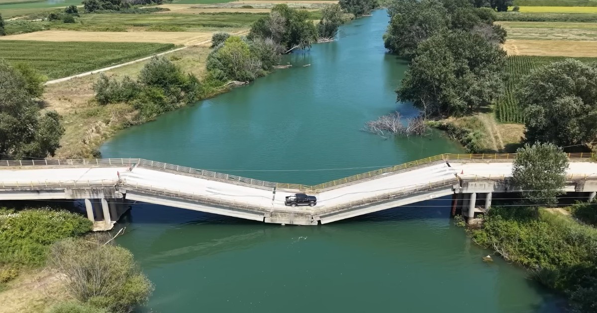 Imaginea prezintă o vedere aeriană a unui pod de beton prăbușit la mijloc peste un râu, cu o mașină neagră în zona surpată.
