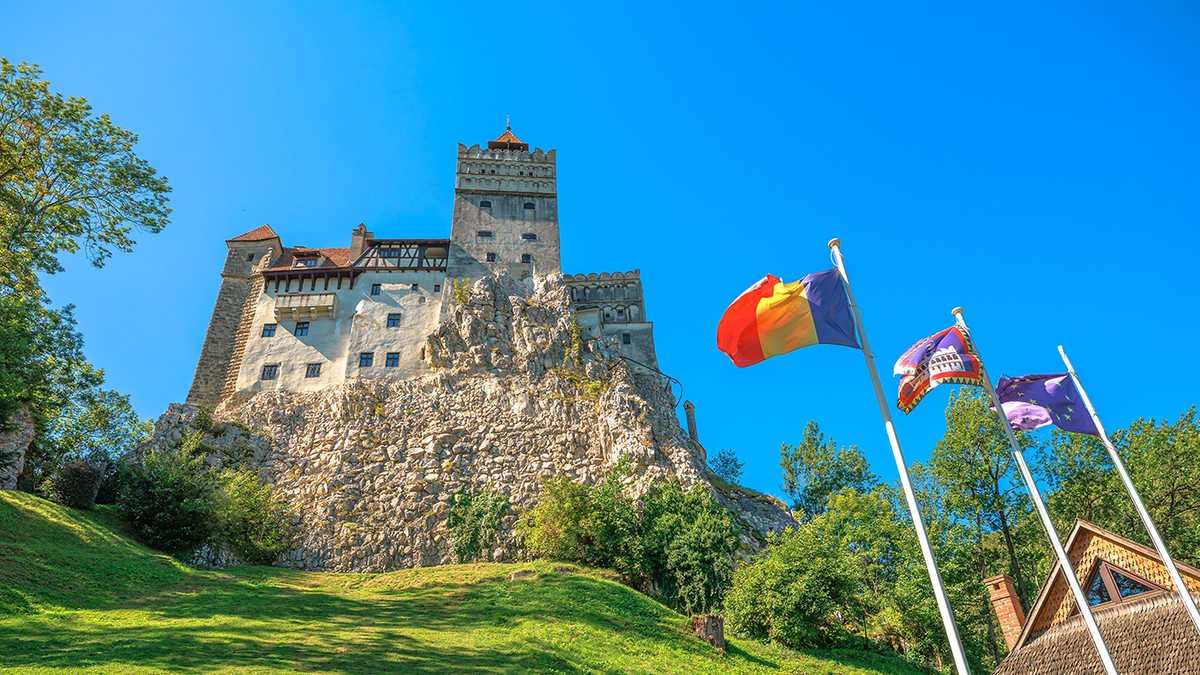 Castelul Bran, aflat pe un deal stâncos, fortăreață medievală și monument național, cu steagurile românești, ale județului Brașov și ale Uniunii Europene fluturând în față, sub un cer albastru senin în România, Transilvania