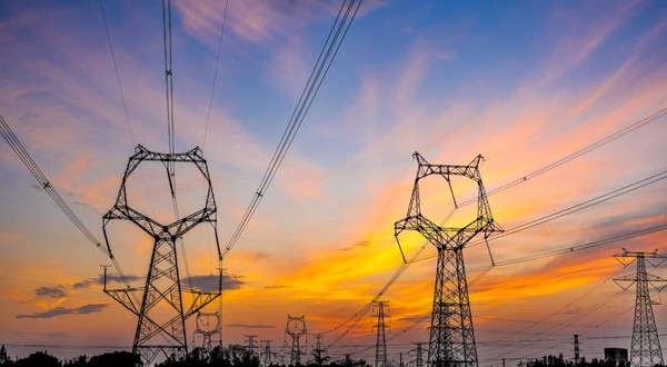 O rețea de energie cu stâlpi de curent de înaltă tensiune și cabluri interconectate, fotografiată într-o zonă de câmpie din România, la apus. Foto: Shutterstock