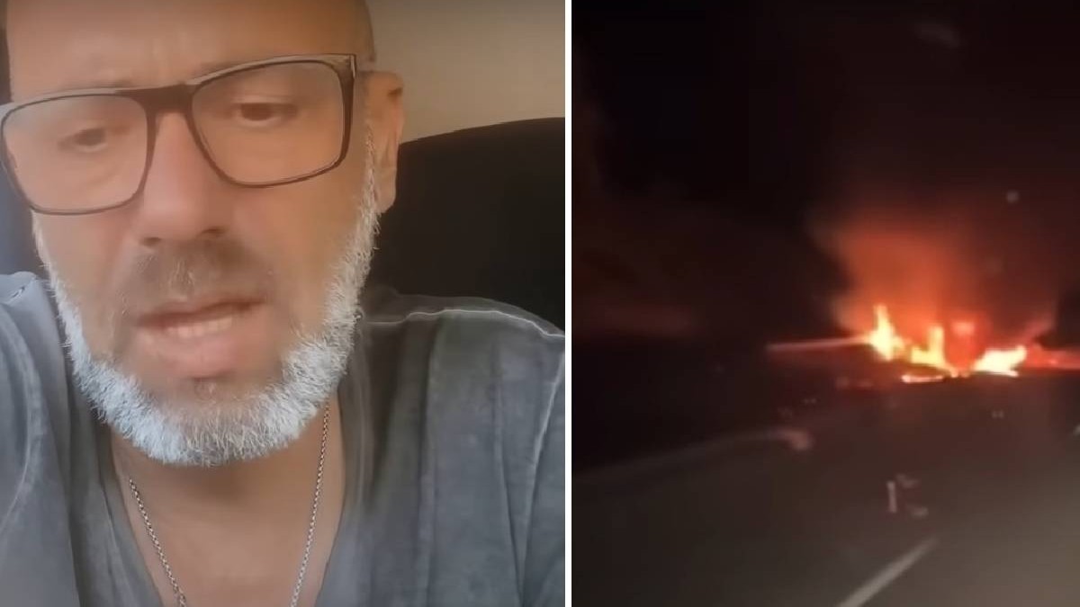 Un bărbat cu ochelari vorbește în fața camerei de luat vederi, în dreapta lui o mașină arde în noapte