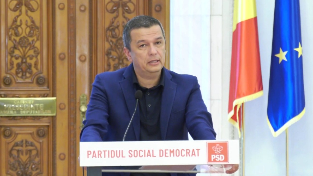 Imaginea îl prezintă pe politicianul Sorin Grindeanu susținând o declarație de la un pupitru inscripționat cu sigla Partidului Social Democrat, având în fundal drapelul României și cel al Uniunii Europene.