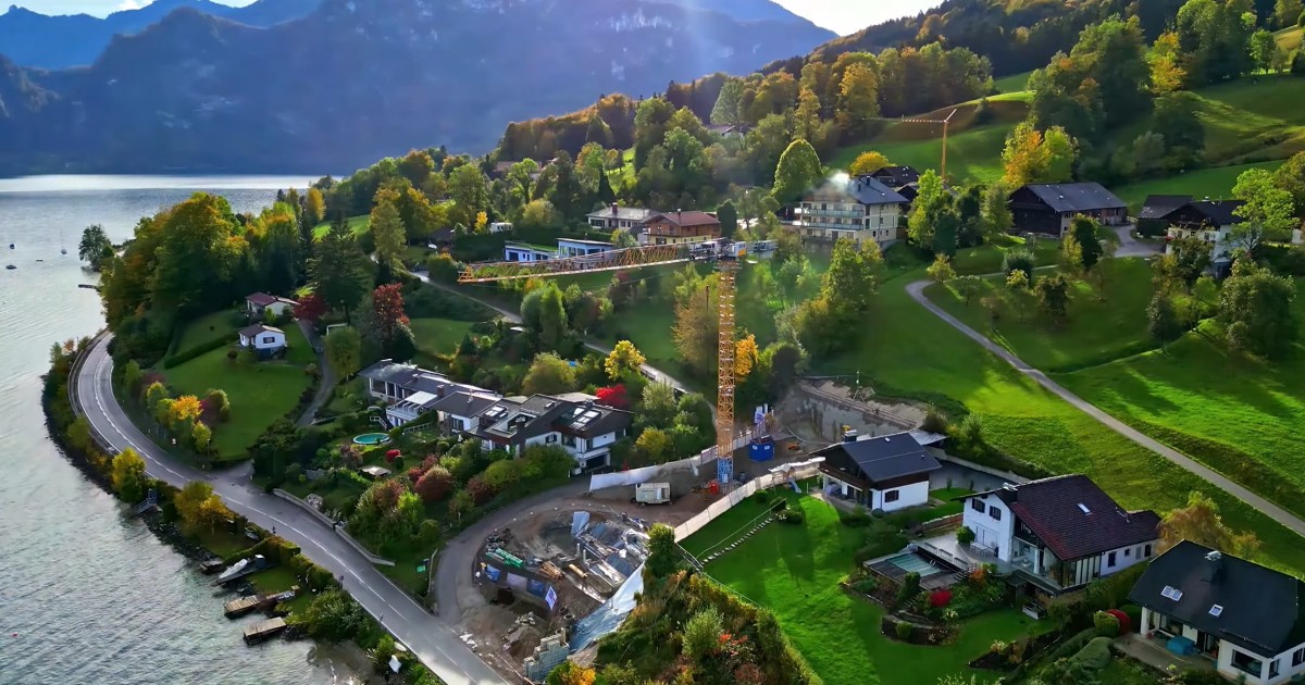 Vedere panoramică aeriană a unui sat situat pe malul unui lac, case cochete și curți verzi dispuse pe un deal împădurit, cu munți în fundal