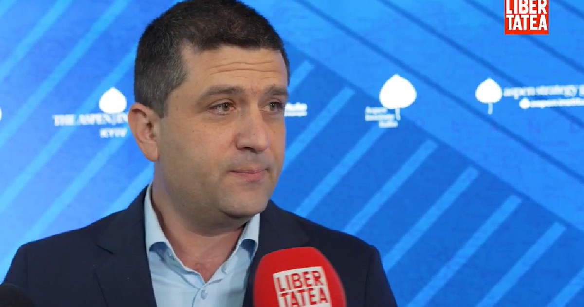 Imaginea îl prezintă pe Radu Miruță, senatorul USR, în timpul unui interviu. El poartă o cămașă albastră deschisă și un sacou bleumarin, iar pe față are o expresie serioasă, privirea fiind îndreptată spre stânga, în afara cadrului. În prim-plan, se vede un microfon roșu, pe care este inscripționat logo-ul ziarului "LIBERTATEA". În fundal, se observă un panou albastru cu diverse logo-uri și inscripții, inclusiv "ASPEN INSTITUTE", sugerând că interviul a avut loc în timpul unui eveniment organizat de această instituție. În colțul din dreapta sus al imaginii este afișat din nou logo-ul "LIBERTATEA".