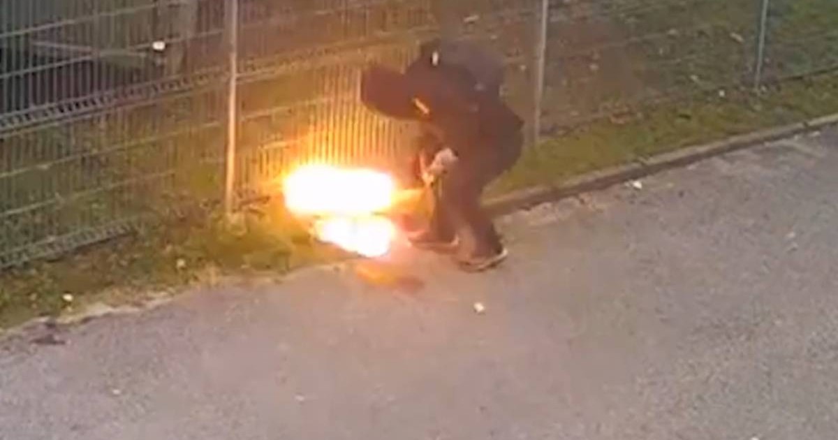 Barbat imbracat in negru complet, cu o vagula pe cap, in timp ce da foc la ceva langa un gard
