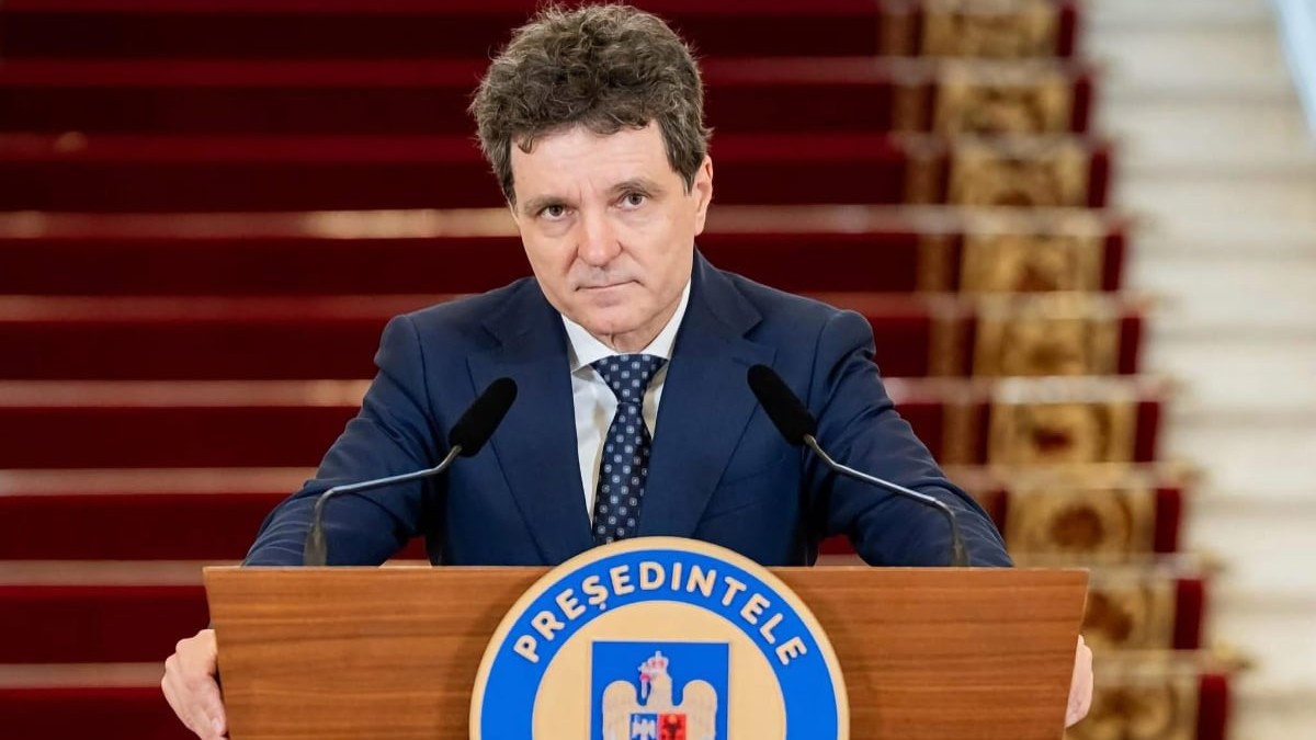 Nicușor Dan îmbrăcat în costum bleumarin și cravată cu model, privind spre cameră din spatele unui pupitru oficial.