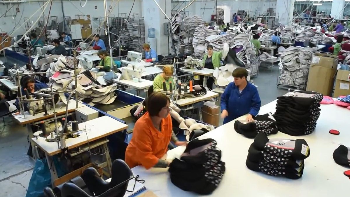 Imagine din interiorul unei fabrici, cu rânduri de mașini de cusut, muncitori în uniforme și grămezi de țesături.