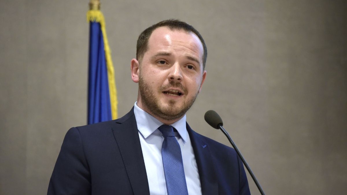 Alexandru Robodete, îmbrăcat într-un costum negru cu o cămașă albă și o cravata albastru închis, vorbind la microfon. în spatele sau se află un drapel al României
