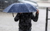 O persoană îmbrăcată într-o geacă neagră care se adăpostește sub o umbrelă albastră, ținând-o strâns cu ambele mâini în timpul unei ploi torențiale.