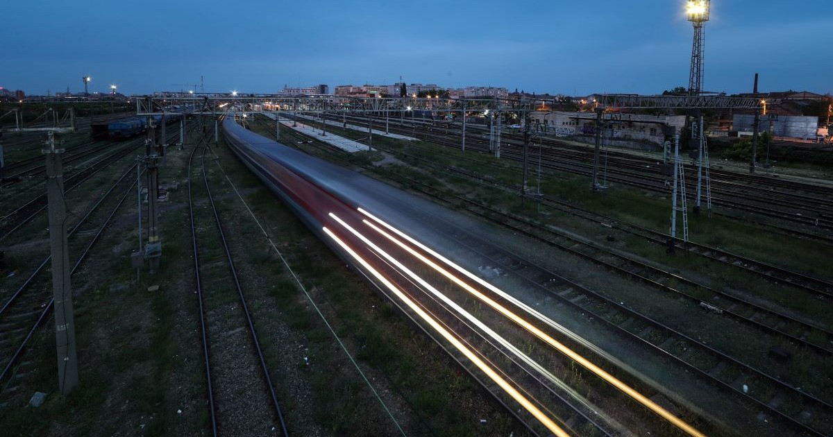 Un tren în mișcare pe timp de seară, lăsând dâre luminoase pe o cale ferată complexă, într-o zonă urbană iluminată