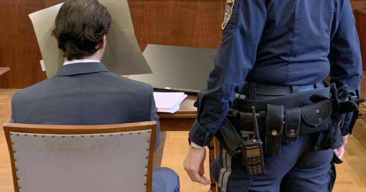 Imaginea arată un bărbat în costum, văzut din spate, stând la masa dintr-o sală de judecată. Își acoperă fața cu un paravan gri. Lângă el stă un polițist în uniformă albastră, cu echipament pe centură.