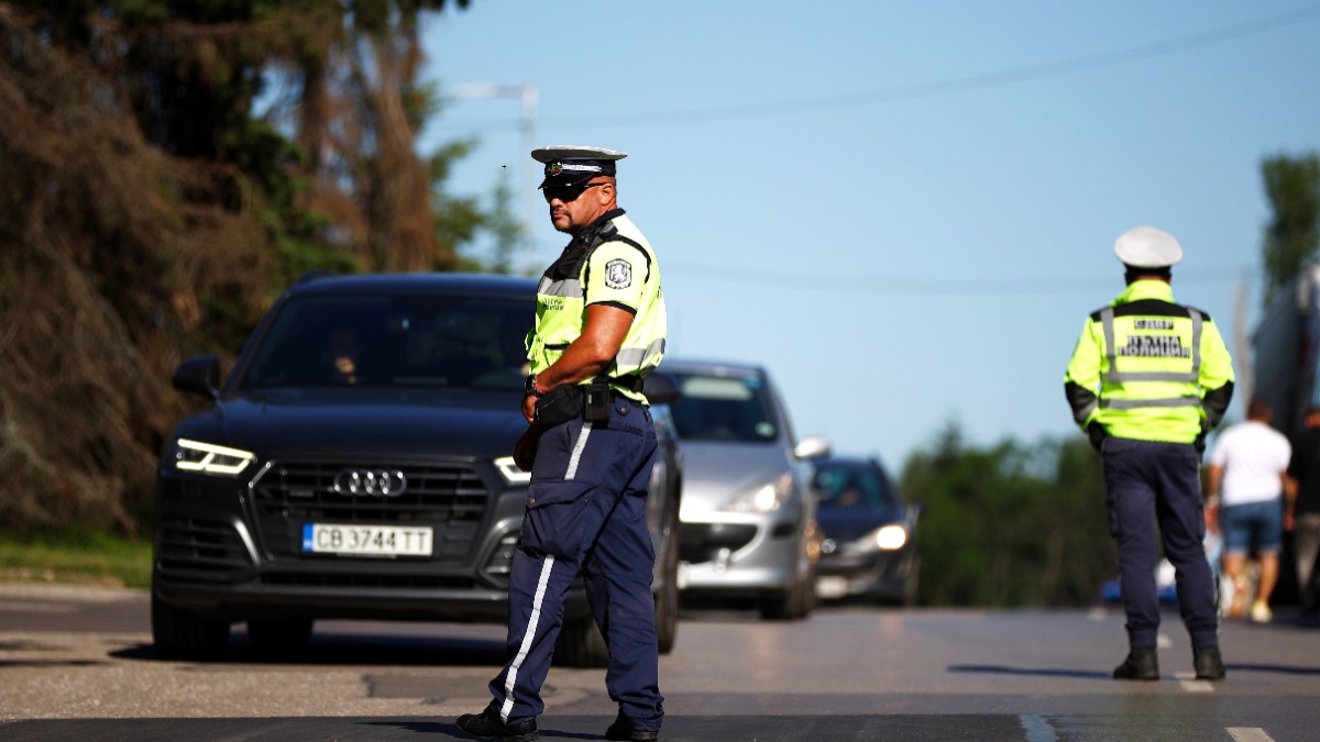 Doi polițiști bulgari de la poliția rutieră, îmbrăcați în veste reflectorizante, supraveghează traficul pe un drum public unde sunt oprite mai multe autovehicule.
