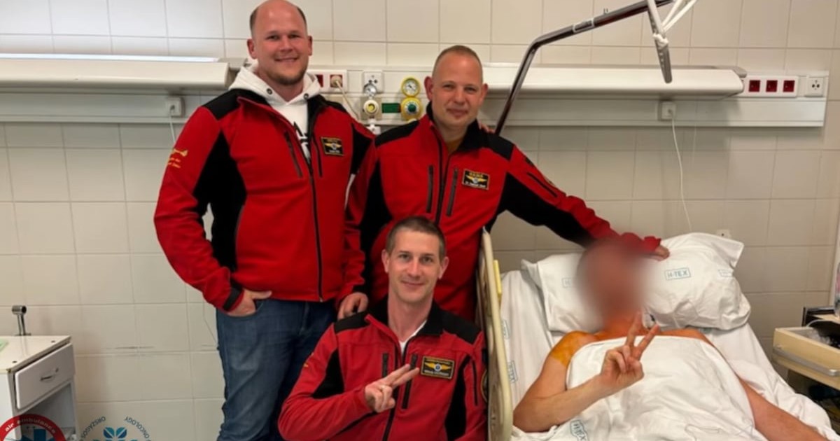 Un bărbat pe patul de spital. Lângă acesta se află trei bărbați în echipament roșu.